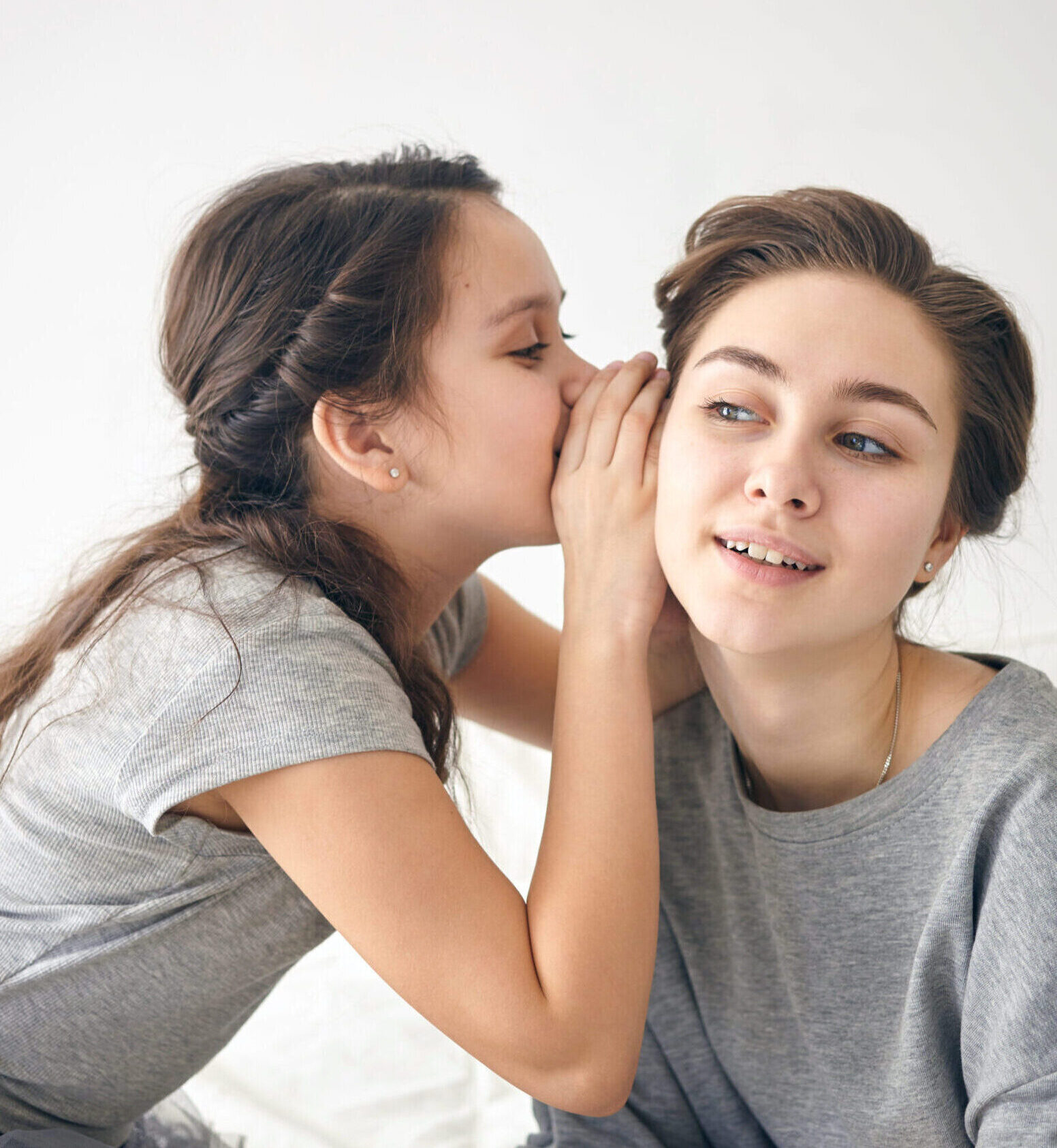 Horizontal picture of beautiful young female having curious interested look while listening to her daughter, who is telling secret, whispering into ear. People, secrecy and body language concept
