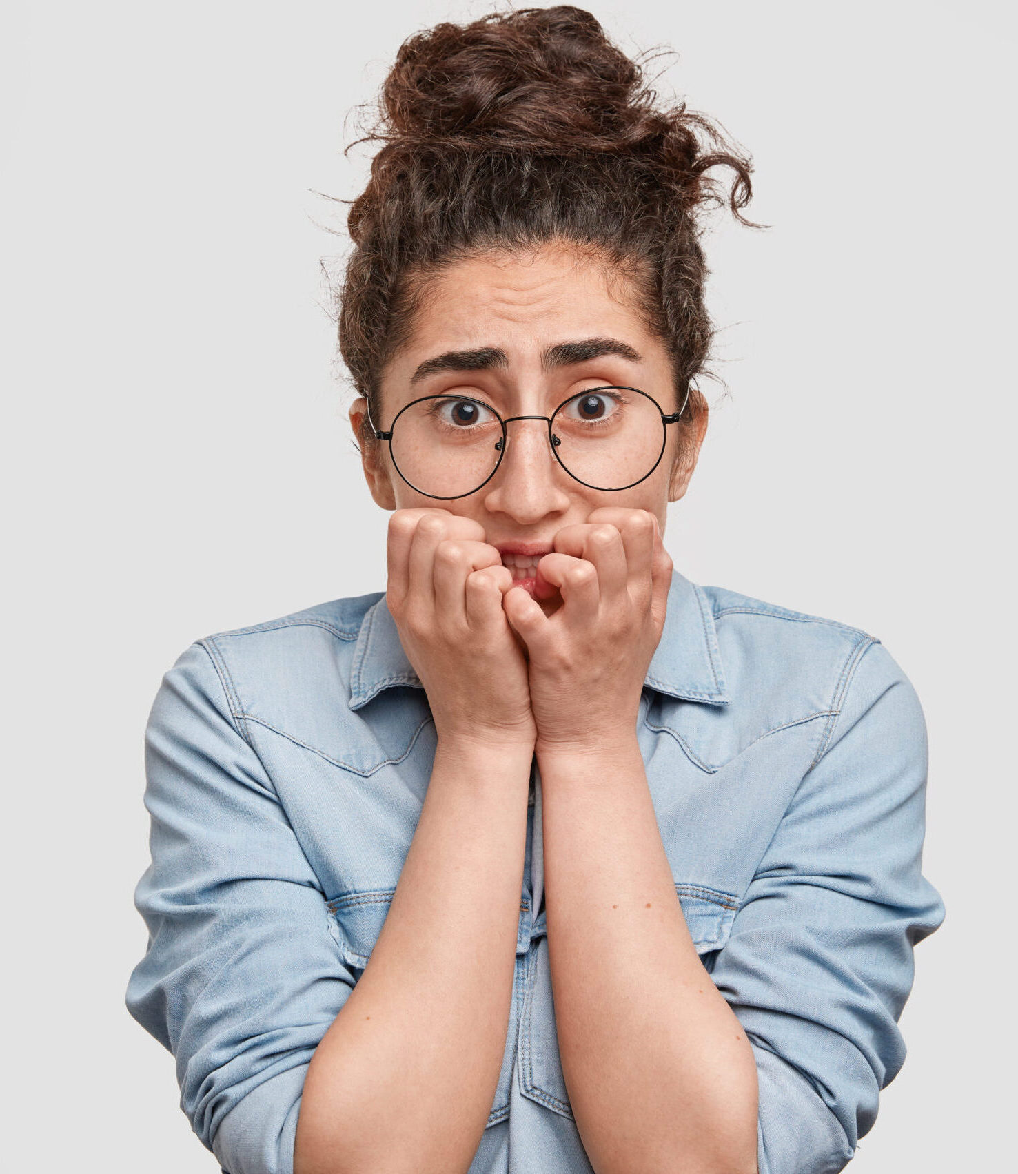 Beautiful young female model bites finger nails and looks with fear at camera, worries if someone knows her secret, dressed in denim jacket, isolated over white background. People and problems