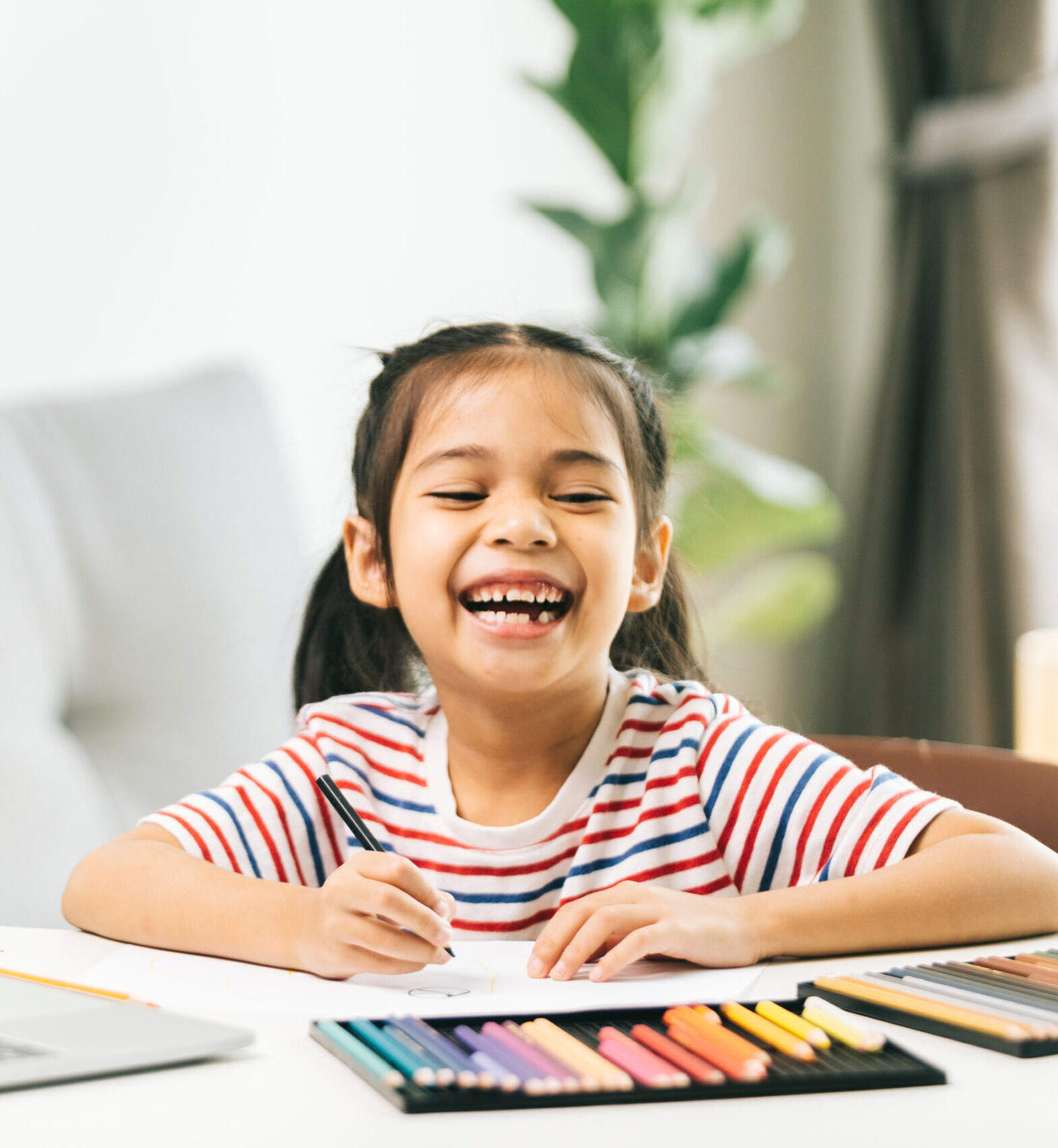 Little asian girl drawing with color pencils while using laptop at home